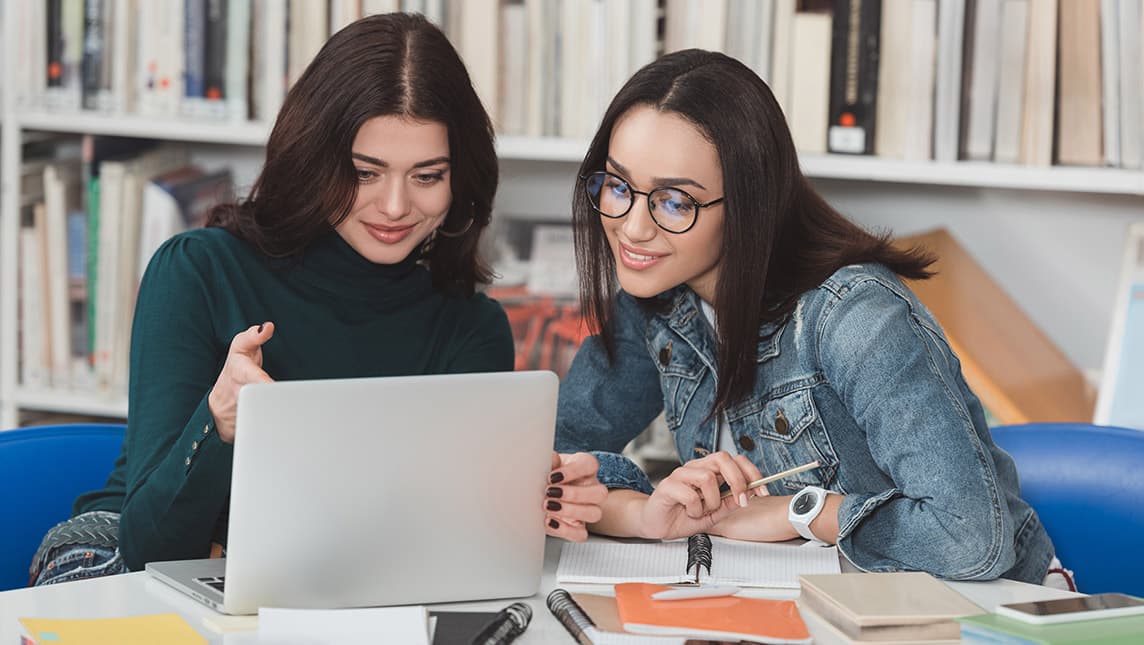 Two students discuss an unseen laptop screen in a library.